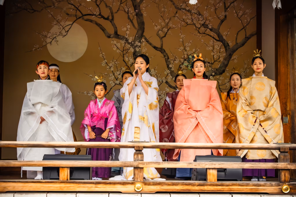 A group of Mikos stand on a kaguraden with a cherry blossom and moon painted background.