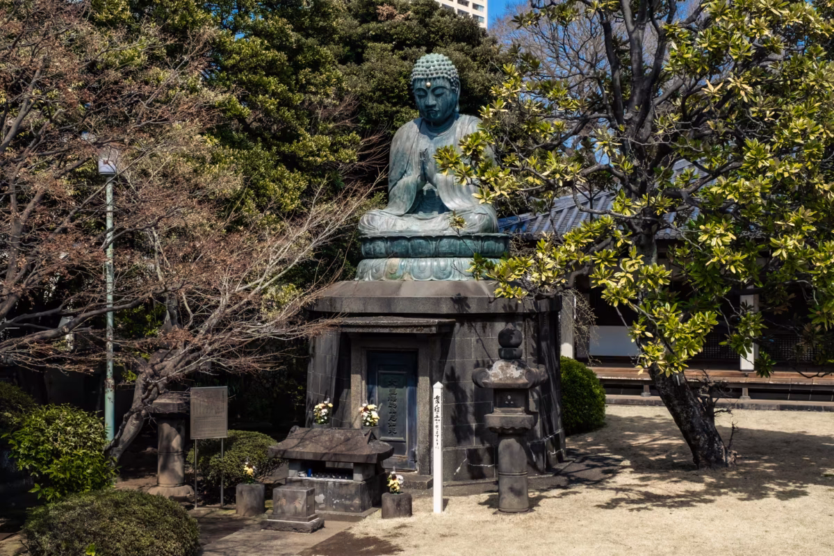 A large Buddha statue sits upon a large pedestal in a temple garden.