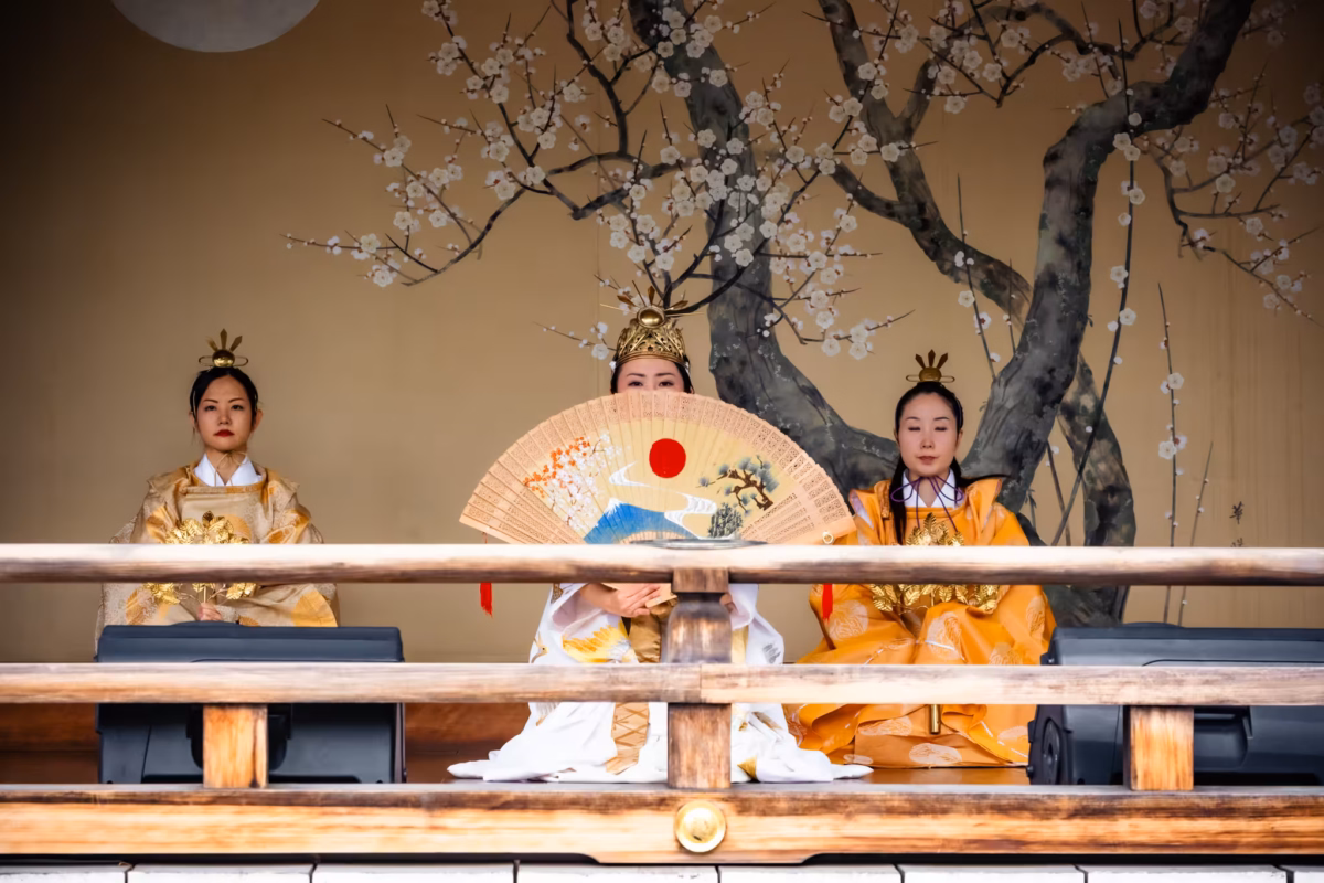 Three mikos kneel on a kaguraden with a painted cherry blossom background.