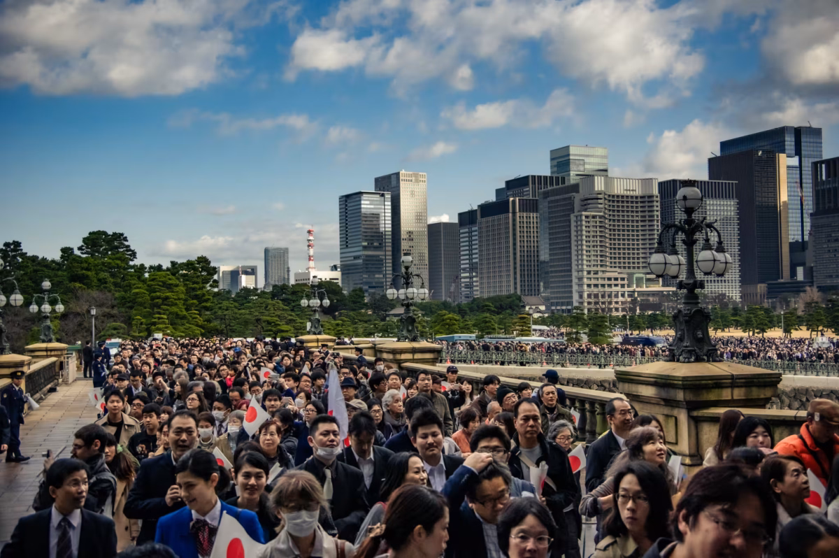 People walk over a stone bridge with skyscrapers in the background.