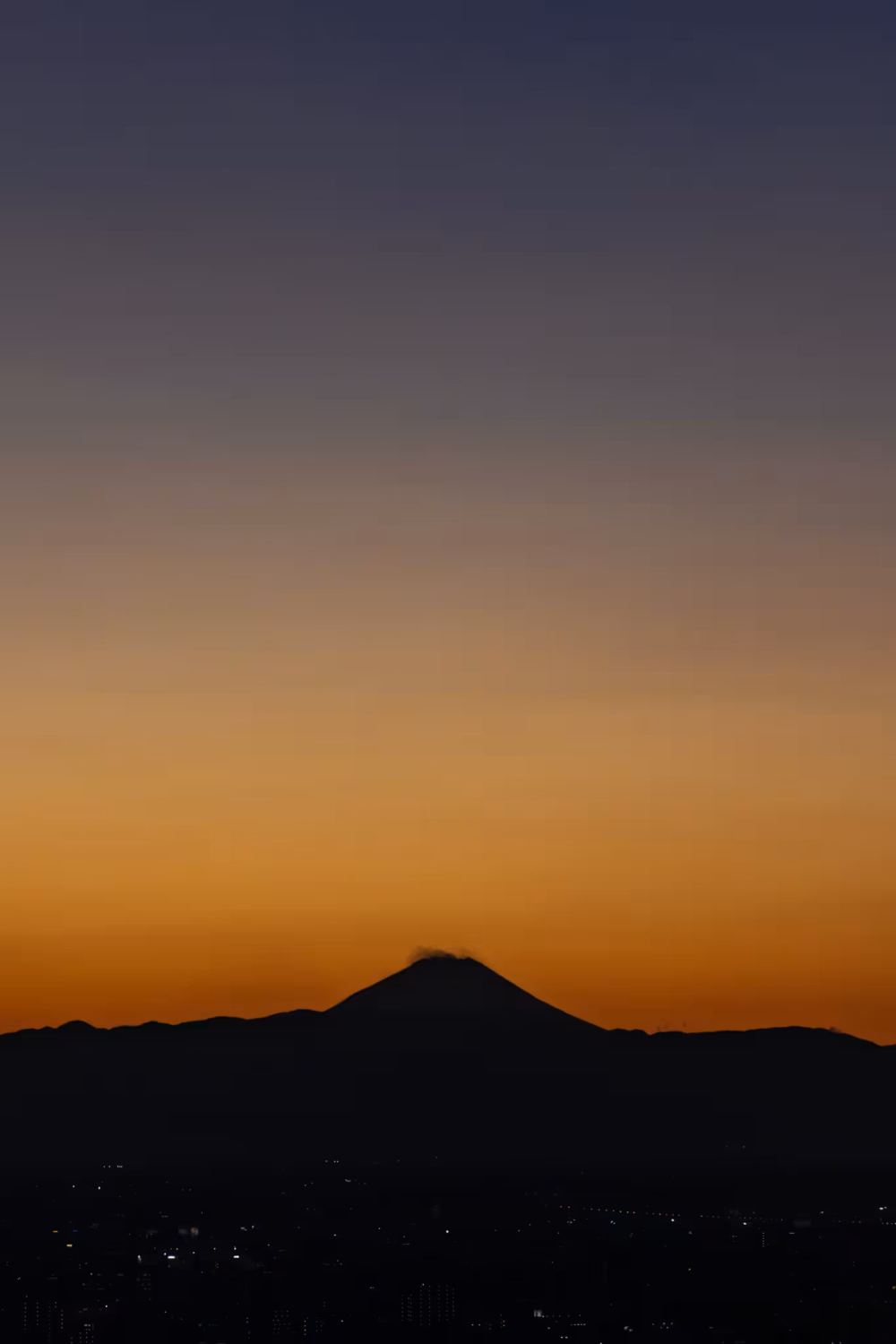 Silhouette of Mount Fuji against a vibrant orange and purple sunset sky over distant, twinkling city lights.