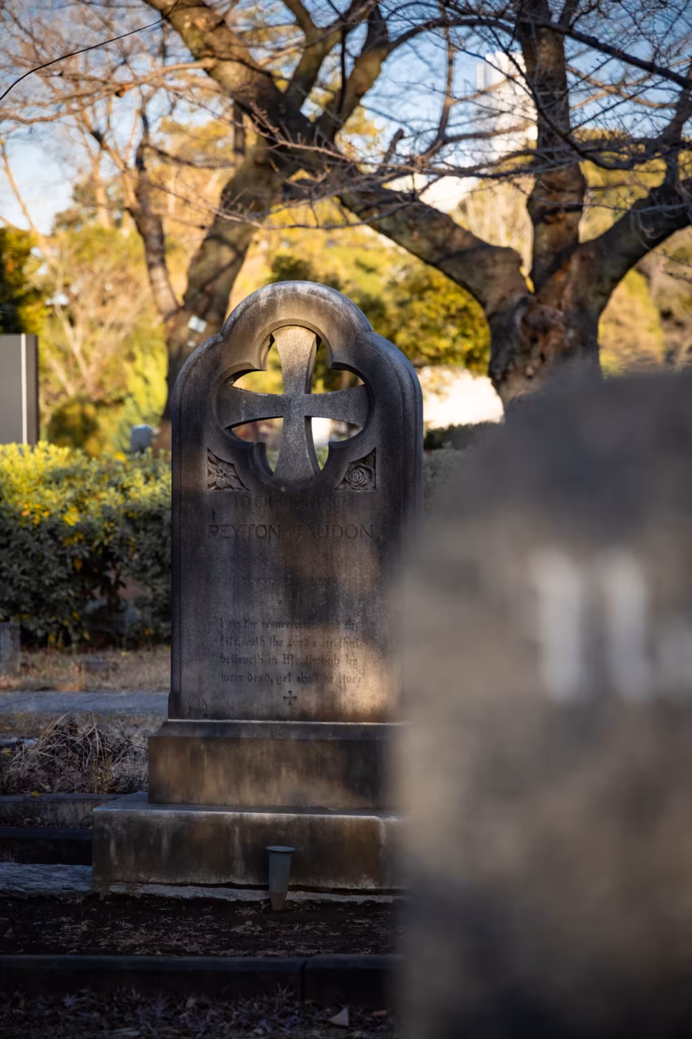 The weathered headstone of Peyton Jaudon with a cross cutout stands in Tokyo’s Aoyama Cemetery during the golden hour.