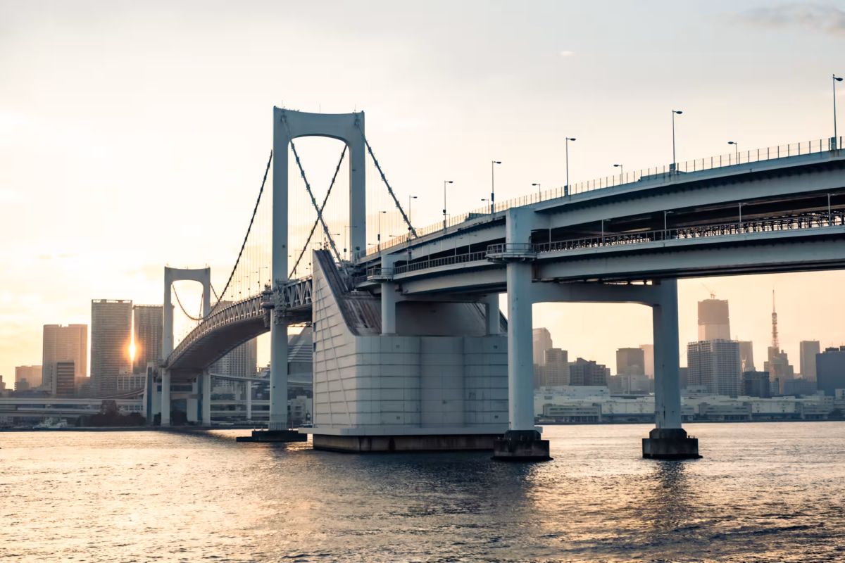 The sun shines between two buildings with Rainbow Bridge in the foreground.