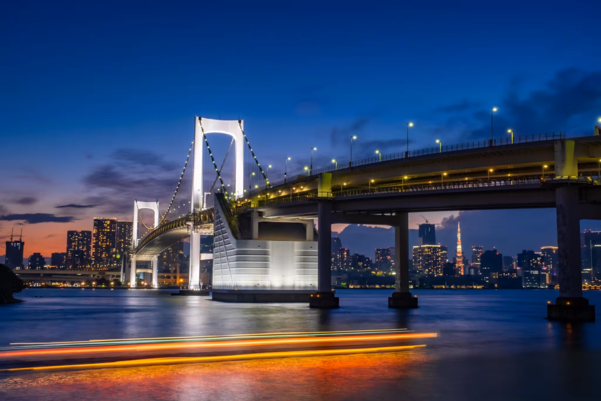 A boat leaves light trails as it passes an illuminated Rainbow Bridge.