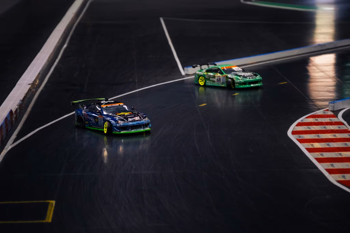 Two RC cars in unison drift around a corner on an indoor tracks.