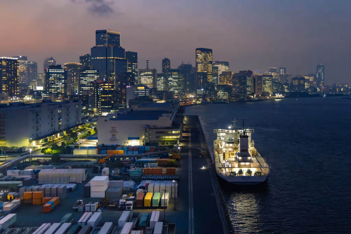 A small ship next to a dock that is crowded with containers.