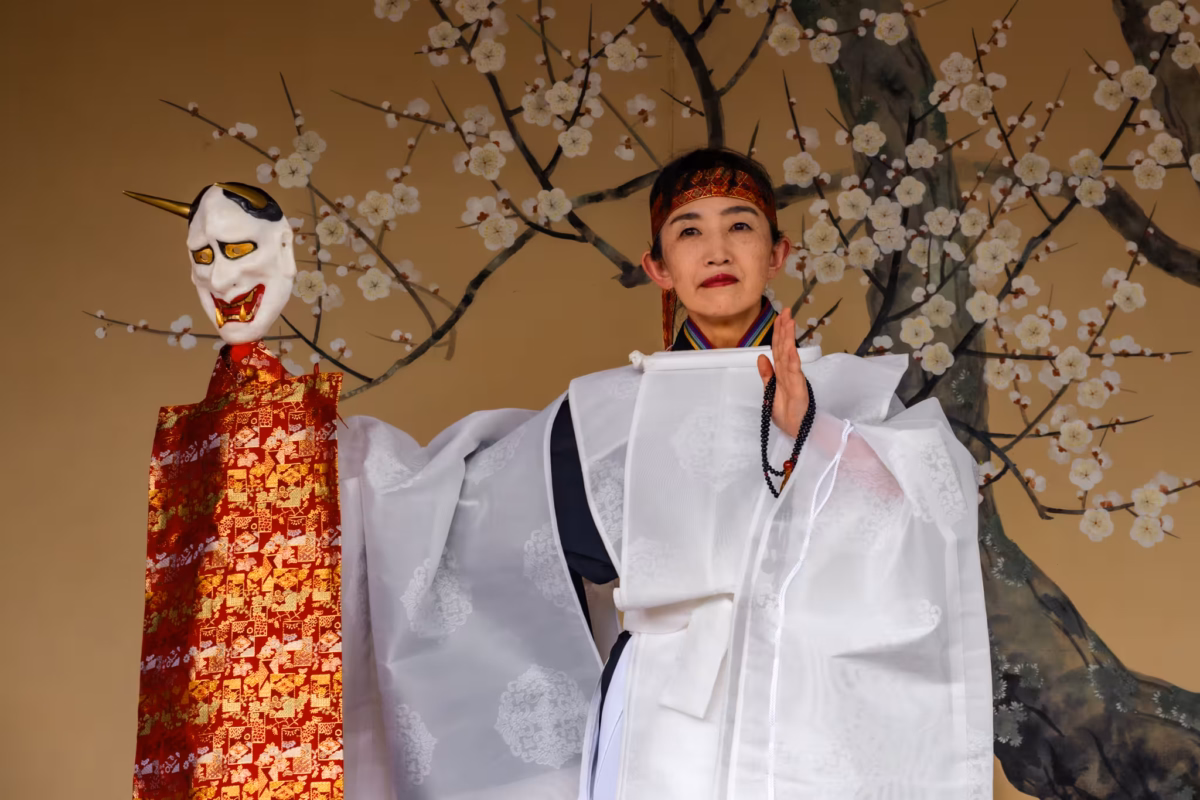 A miko in a white hakui holds a demon mask.