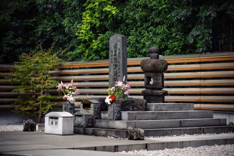A grave and lantern sit in front of a fence and greenery.