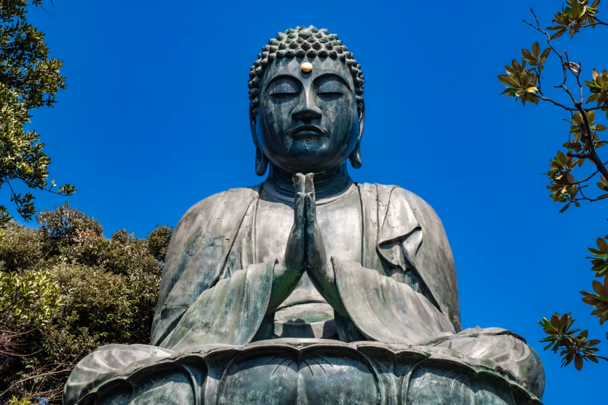 The Genroku Daibutsu sits atop a pedestal in a prayer pose against a blue sky.