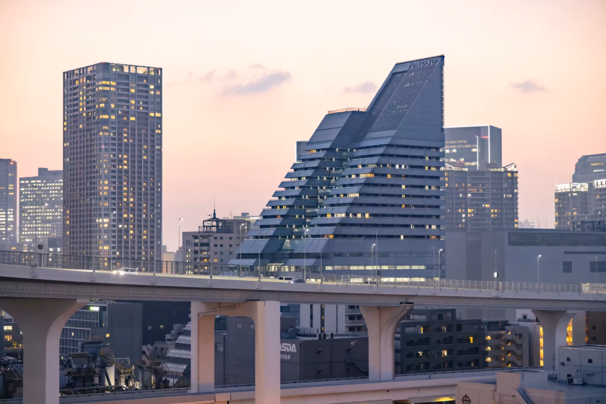 Yokoso Rainbow Tower sits among a large buildings and a freeway at sunset.