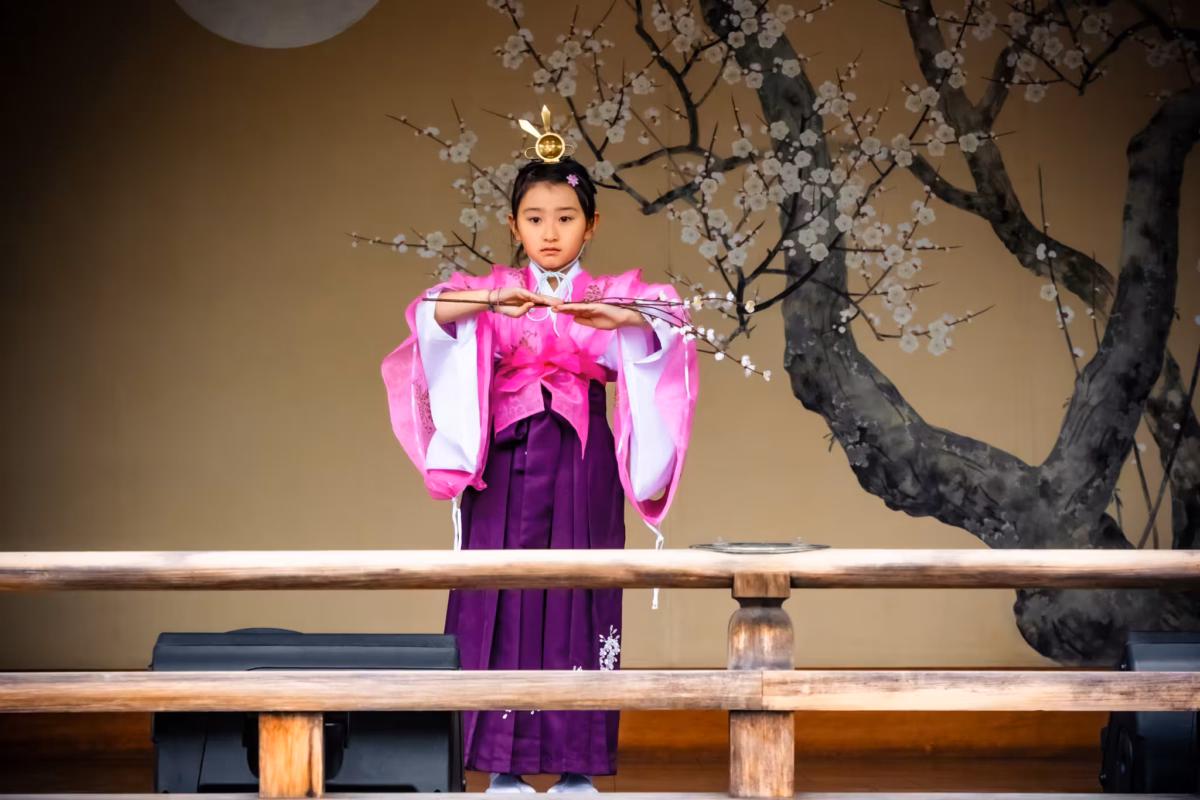 A miko in a pink kimono is on a stage holding a branch with flowers.