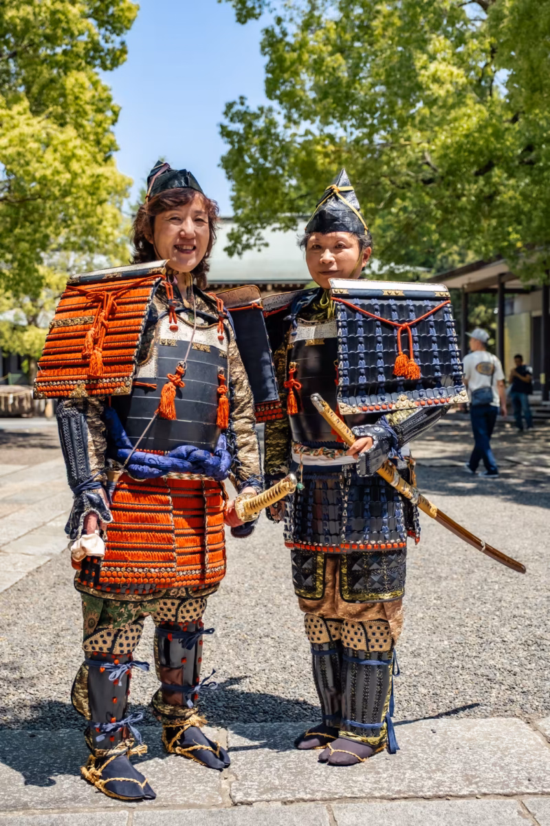 Two female samurai reenactors in Heian-period armor stand with trees in the background.