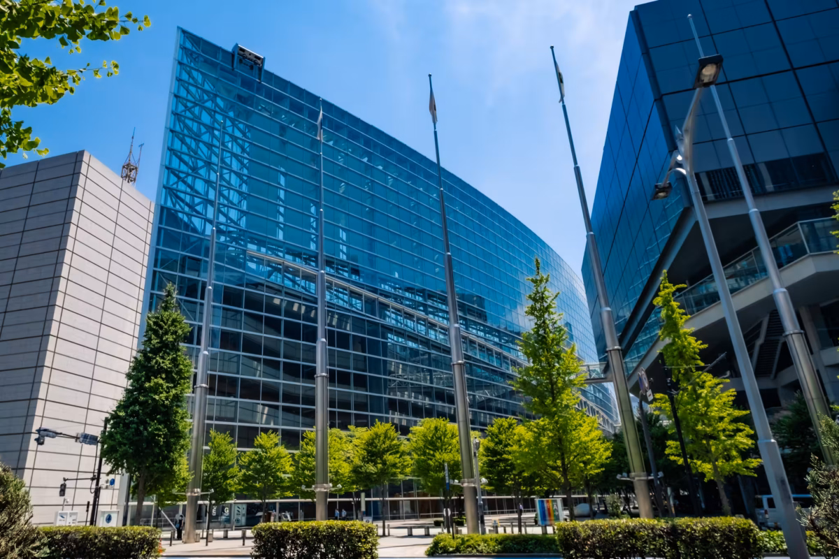 Sleek glass exterior of the Tokyo International Forum with green trees and flagpoles against a clear blue sky.