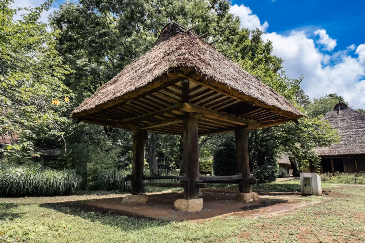 Traditional thatched-roof wooden granary nestled in a lush garden under a bright, cloud-filled blue sky.