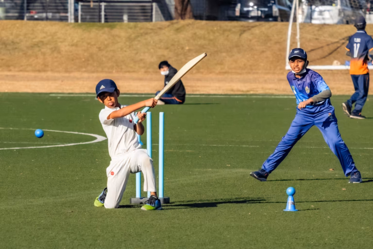 A boy in white swings a cricket bat from a kneeling stance as a teammate in blue prepares to field.