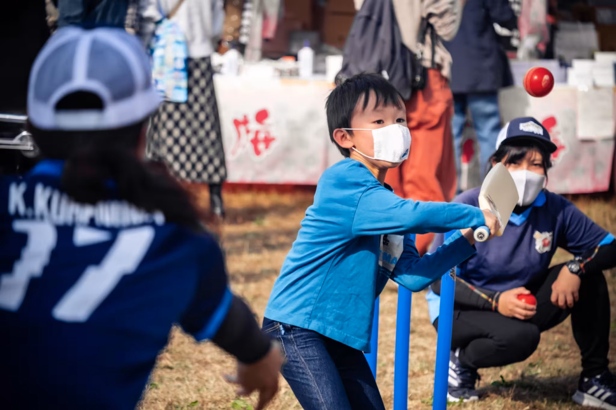 A masked boy swings a plastic cricket bat at a red ball during a lesson at an outdoor festival.