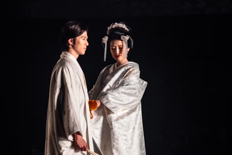 A Japanese fashion model in a white kimono and a headpiece poses with a male model on a stage at a Japanese fashion show.