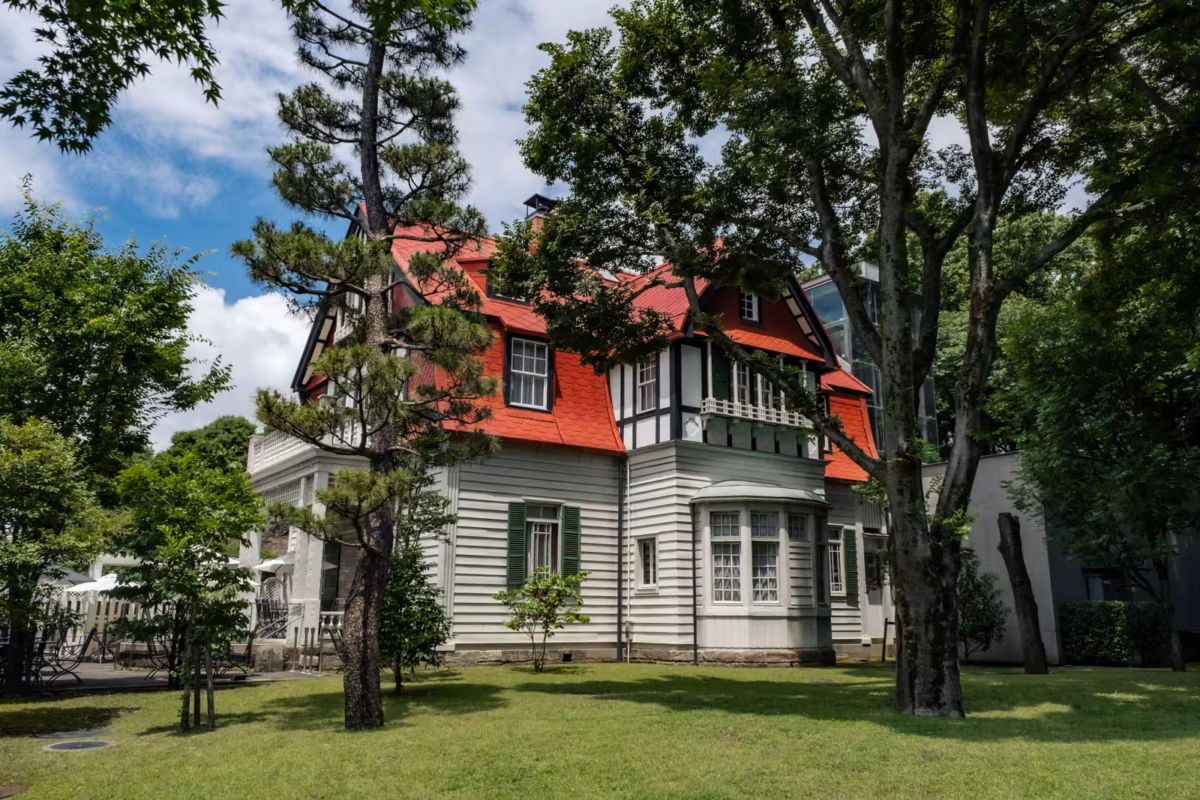 Historic white house with a striking red roof and green shutters, set on a manicured lawn among tall trees.