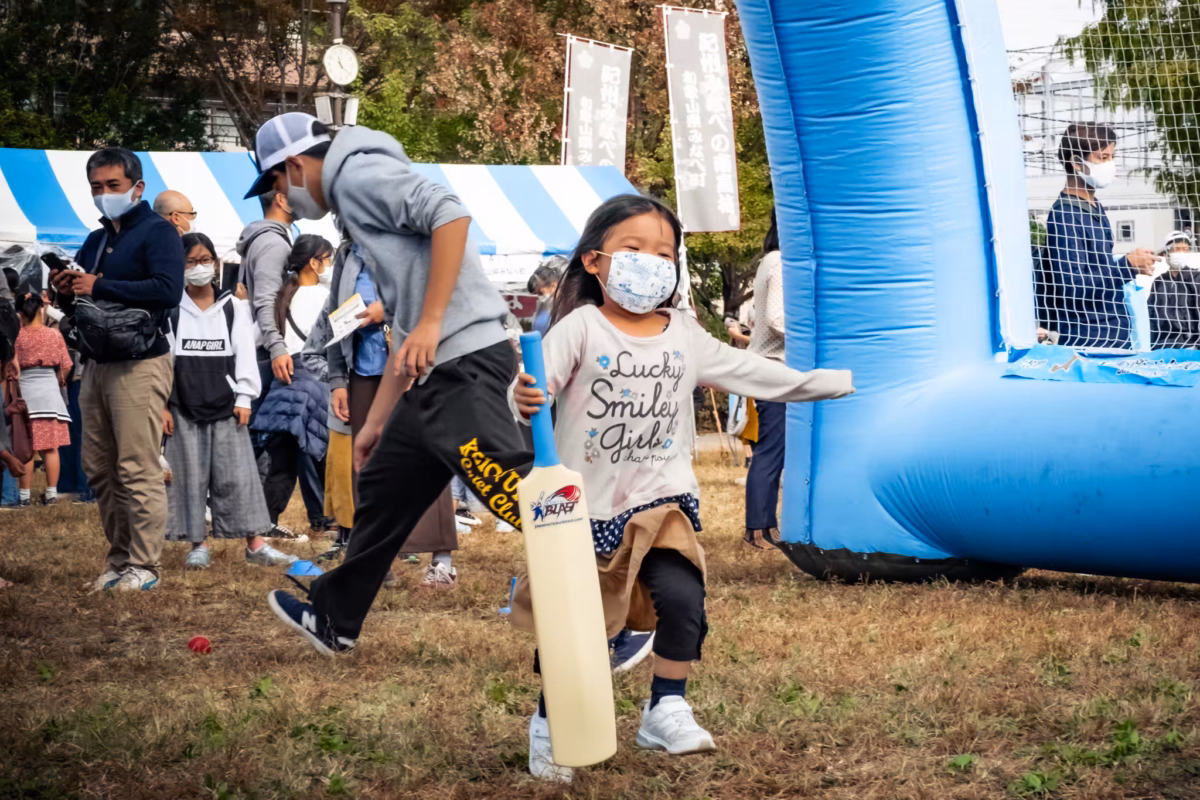 A masked young girl carries a plastic cricket bat past a blue inflatable net at a busy outdoor festival.