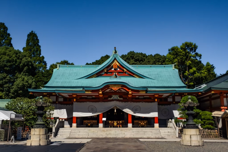 The main hall of Hie Shrine in Tokyo featuring its green copper roof, red pillars, and bronze lanterns under a clear blue sky.