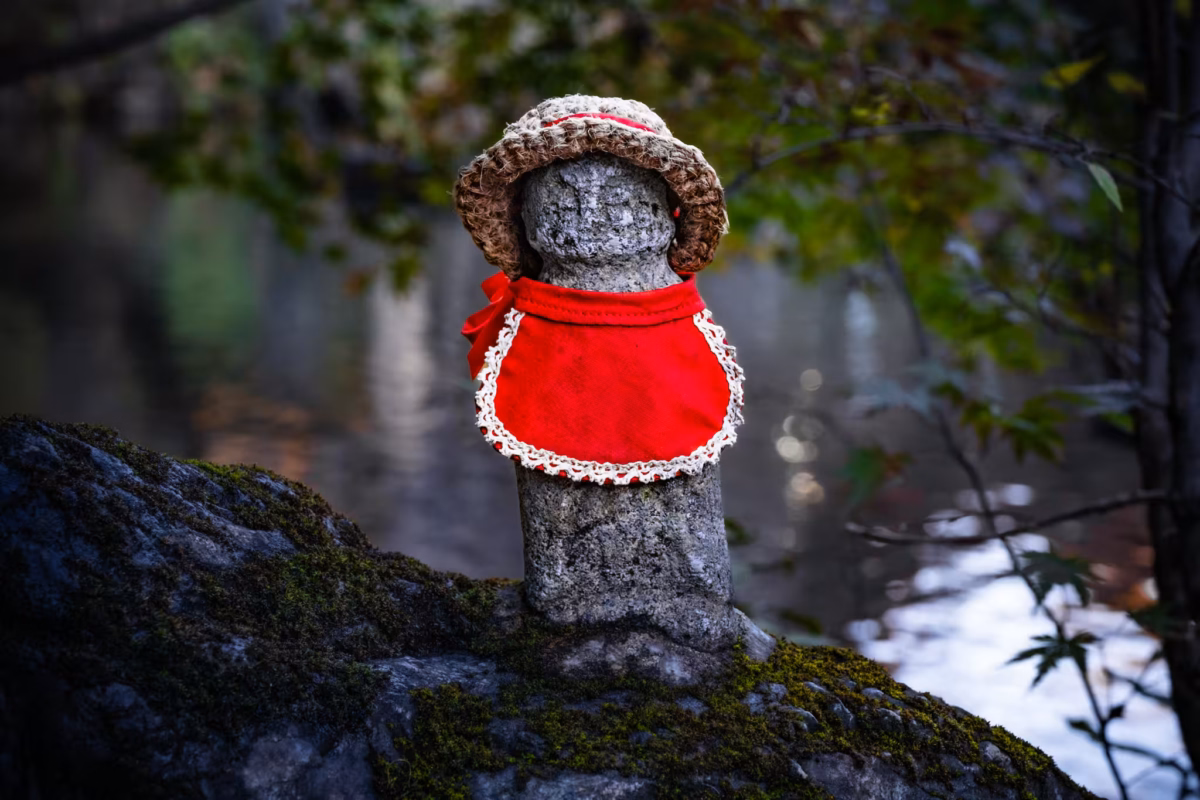 A tiny jizo statue in a red bib and knitted hat sits on a rock.