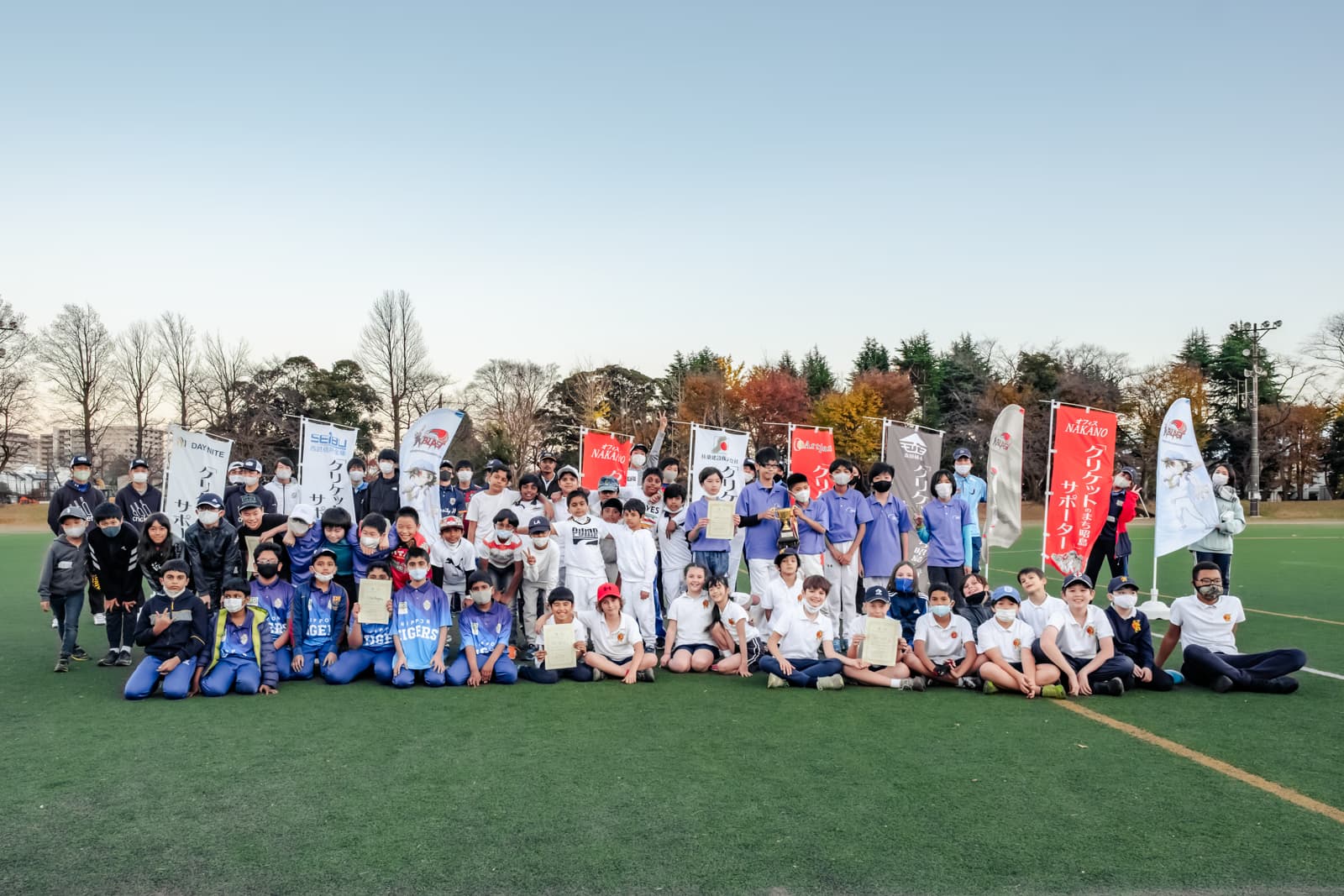 A large group of children and coaches pose with awards and banners at a Japan Cricket Blast event on a green field.