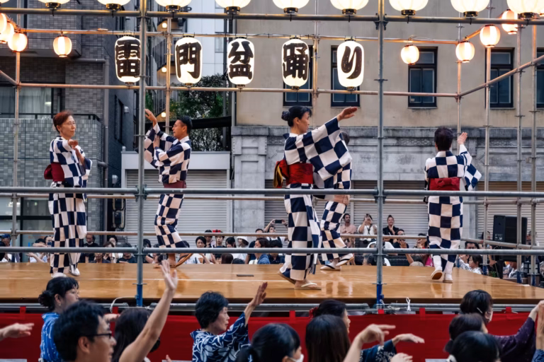 Dancers in checkered yukata perform on a lantern-lit stage at a Japanese Bon Odori festival as the crowd joins in.