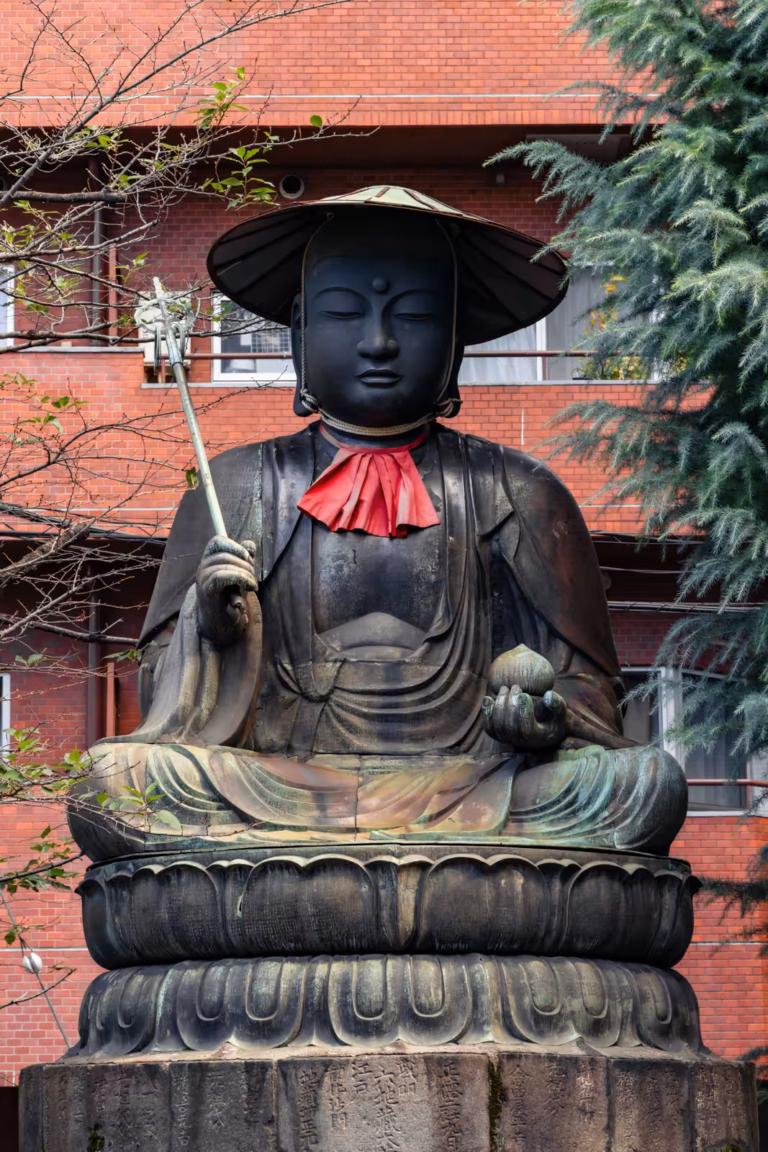 Seated bronze Jizo statue with a red bib and wide hat, holding a staff and jewel, set against a red brick building.