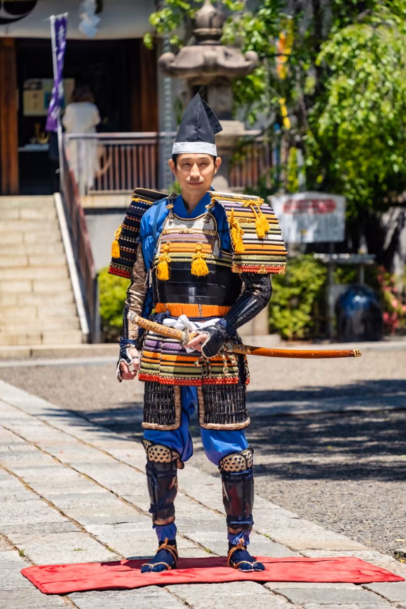 A samurai reenactor in armor and with sword stands on a red mat for photos.