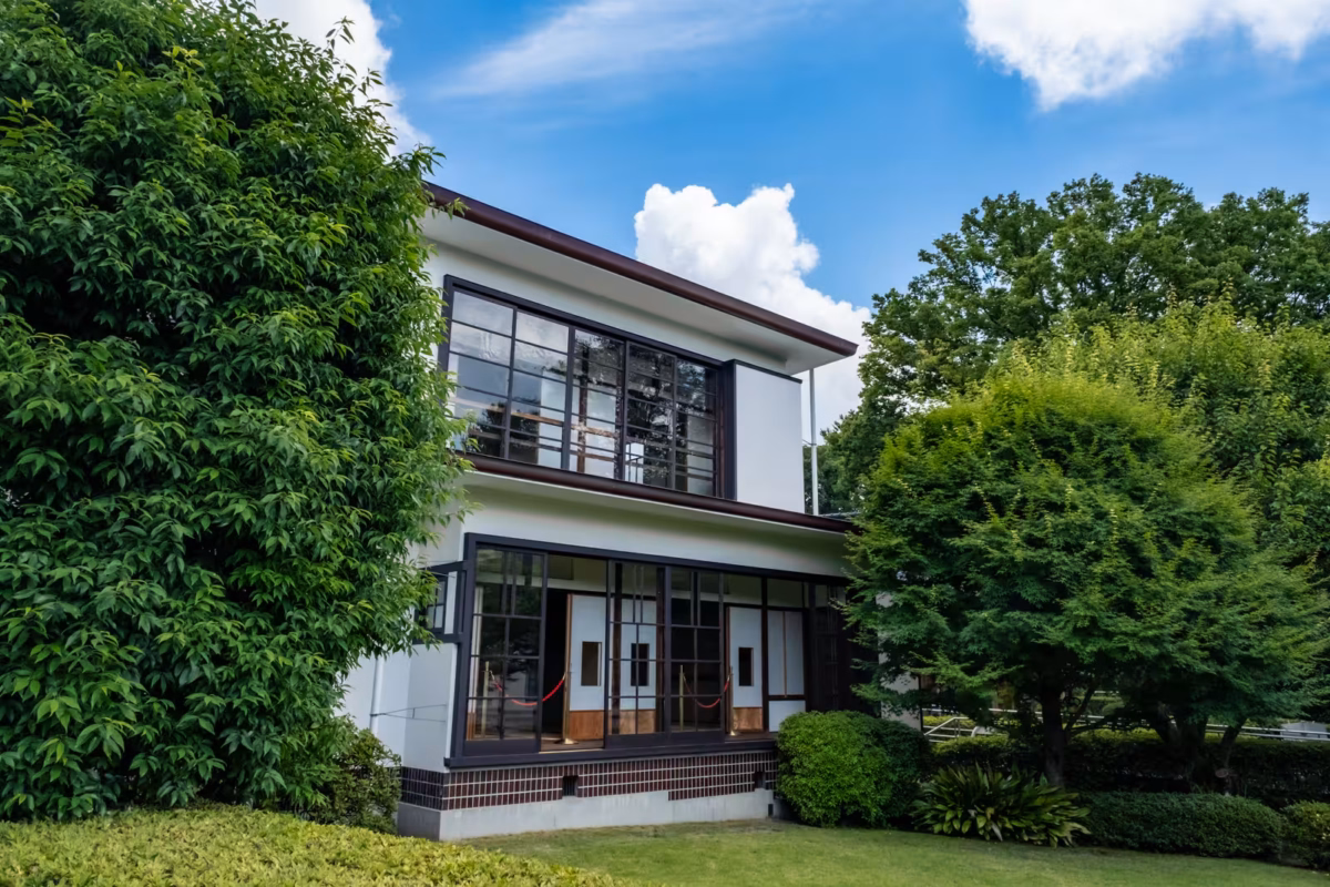 Two-story white house with large glass windows and sliding doors, nestled in a lush garden under a sunny blue sky.