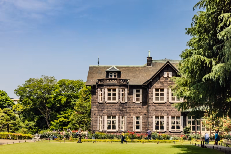 Grand stone mansion at Kyu-Furukawa Gardens with blooming rose beds and visitors on a bright, sunny day.