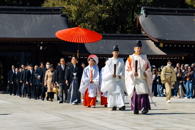 Shinto priests lead a wedding procession with a red parasol across a stone courtyard at Meiji Jingu.