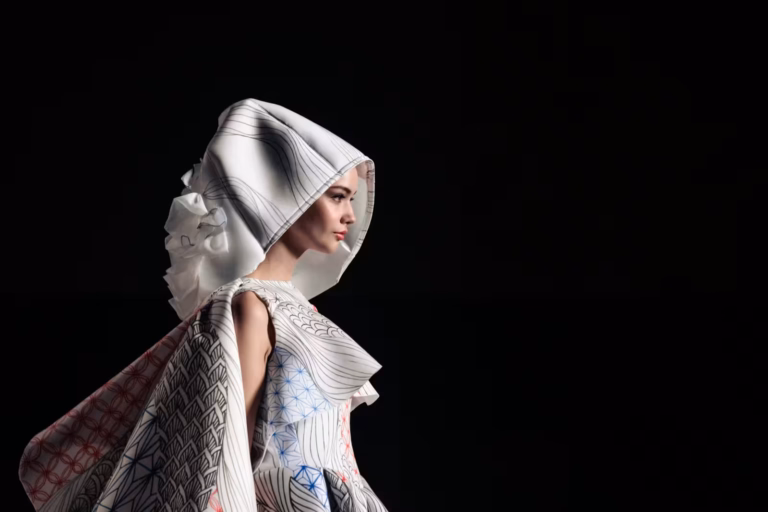 Avant-garde fashion model in a Japanese-influenced white hooded dress with geometric patterns, profile against a black background.