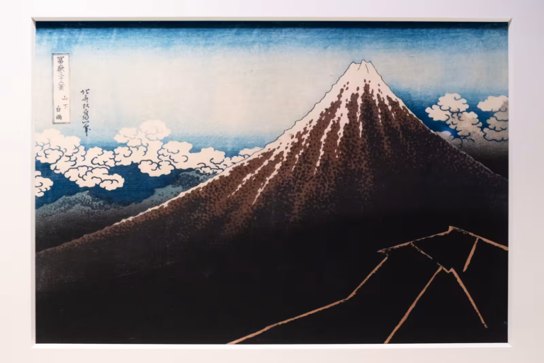 A Japanese woodblock print of Mount Fuji with a dark slope and snowy peak against a blue sky with stylized white clouds.