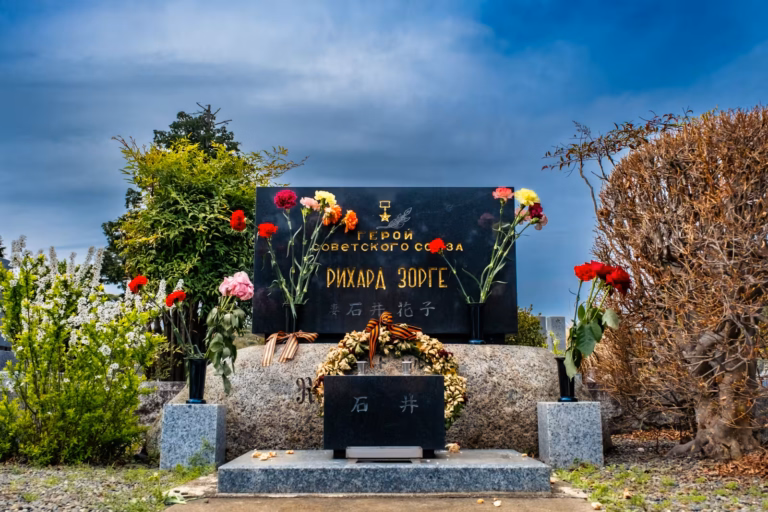 Richard Sorge's grave in Tokyo, featuring a black headstone with Russian text, memorial wreaths, and fresh flowers.