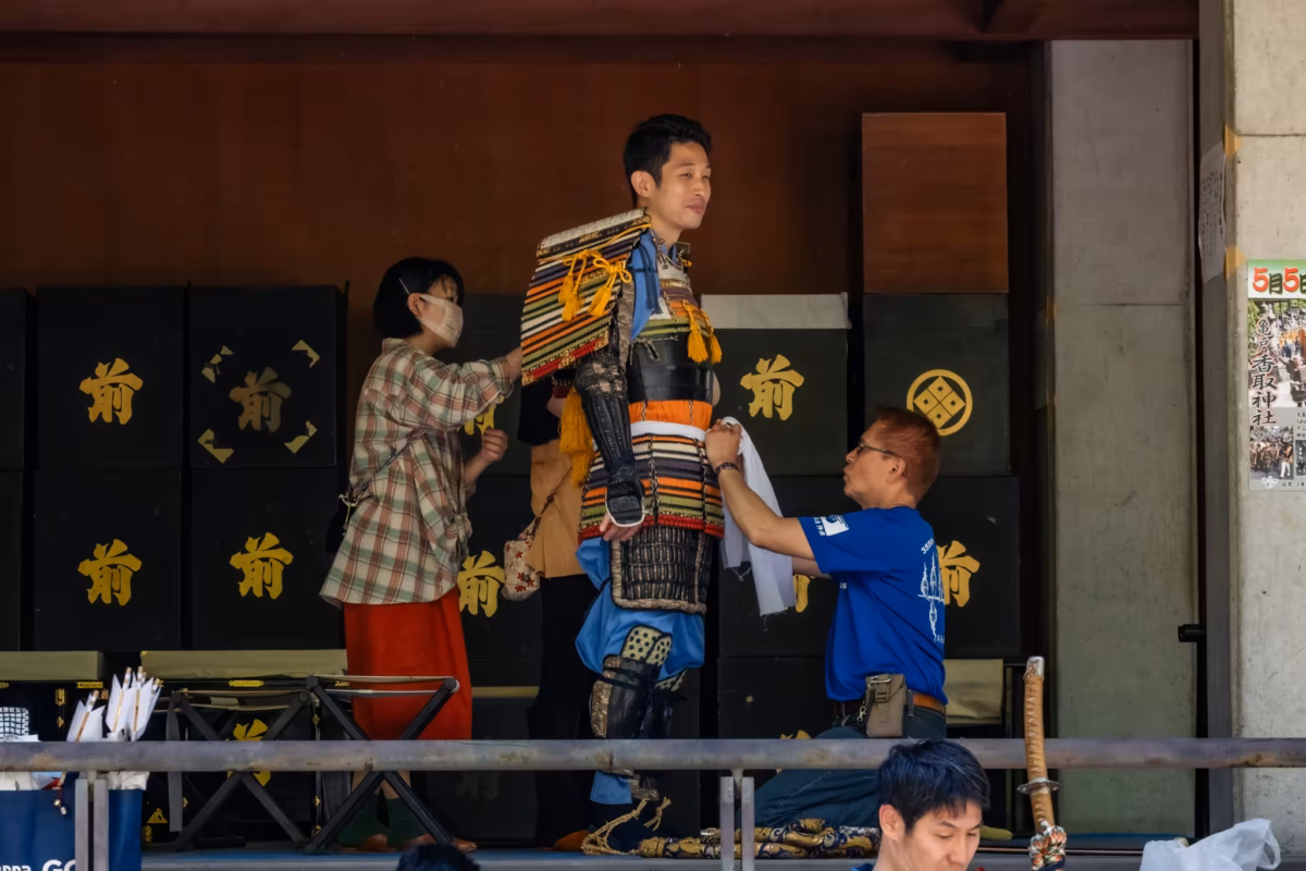 A man and woman assist a man donning samurai for a parade.