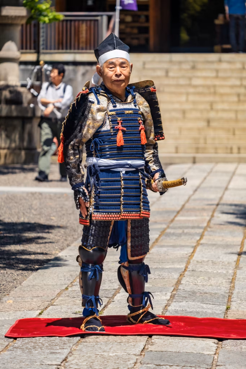 An samurai cosplayer in Heian-period armor stands on a red mat for photos.