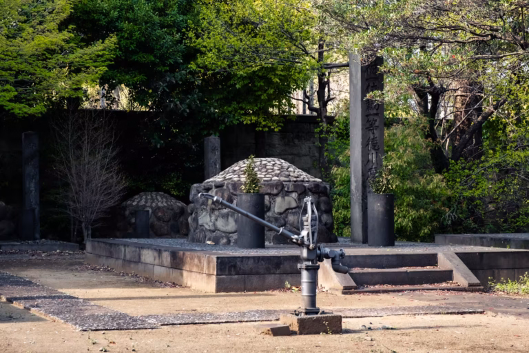 Old water pump and Tokugawa Yoshinobu’s stone cairn tomb at Yanaka Cemetery in Tokyo, surrounded by lush greenery.