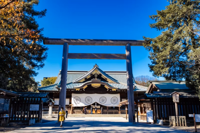 A massive wooden torii gate frames the traditional main hall of Tokyo's Yasukuni Shrine under a brilliant blue sky.