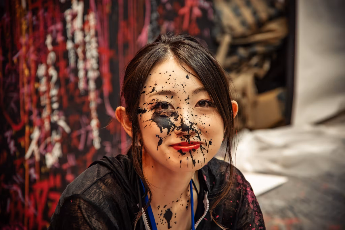 A Japanese woman with paint splashed over her face smiles at the camera.
