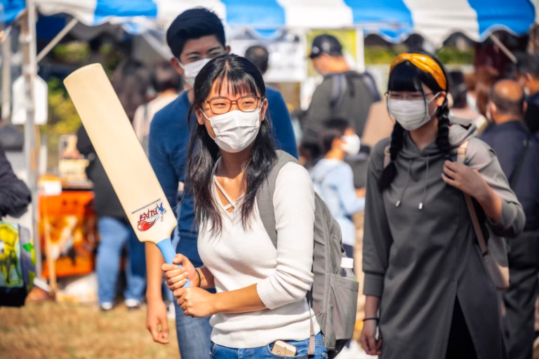 A masked young woman smiling as she holds a cricket bat at a bustling outdoor festival.