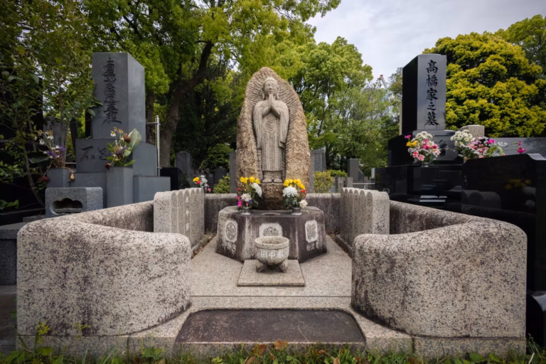 Wide-angle view of a stone Buddha statue grave with fresh flower offerings and modern headstones in Aoyama Cemetery.