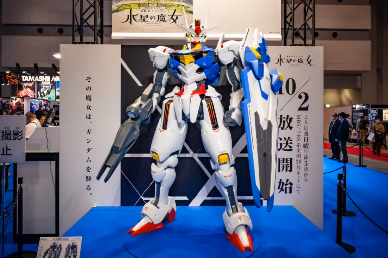 Large Gundam Aerial statue in white, blue, and red with a shield and beam rifle on display at an exhibition booth.