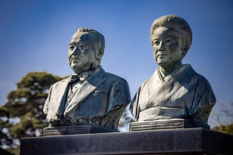 Bronze busts of a man and a woman in kimono at Tama Cemetery, set against a clear blue sky.