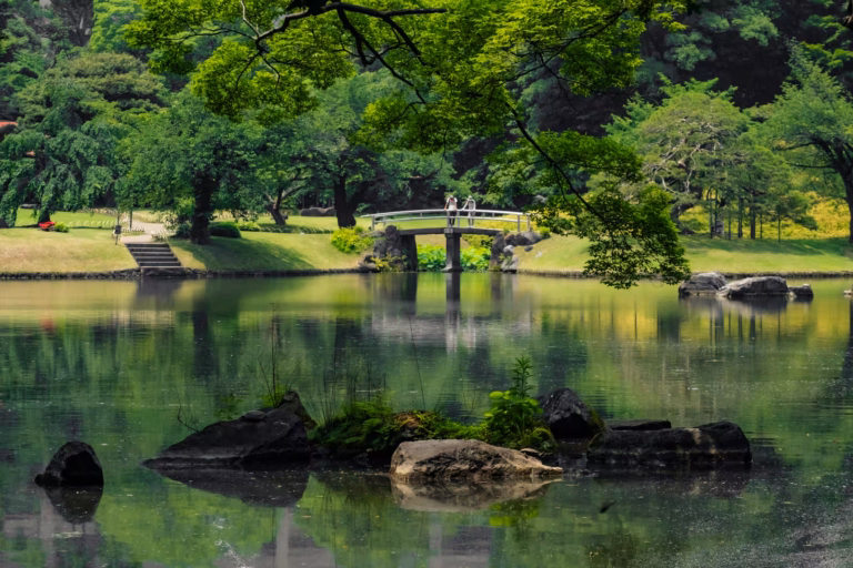 A calm pond at Koishikawa Korakuen with green tree reflections, a white bridge with people, and small rocky islands.