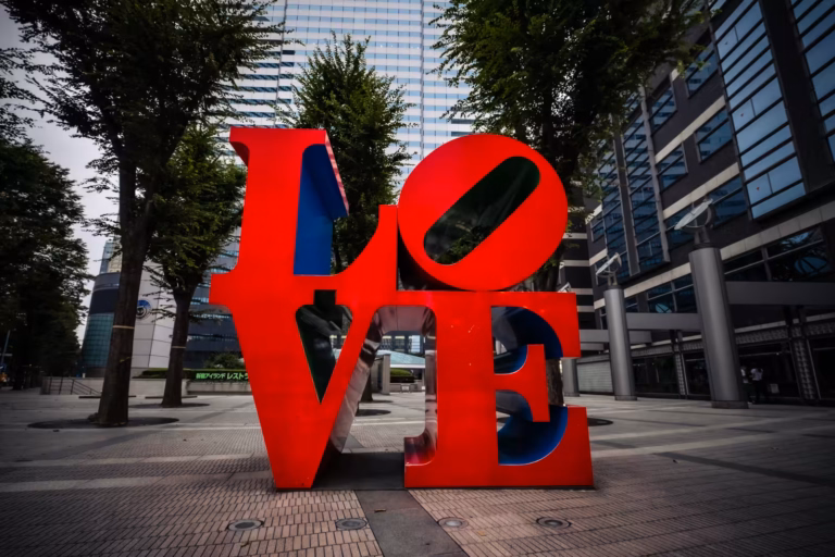 Large red LOVE sculpture centered in a city plaza, framed by leafy trees and tall modern buildings.