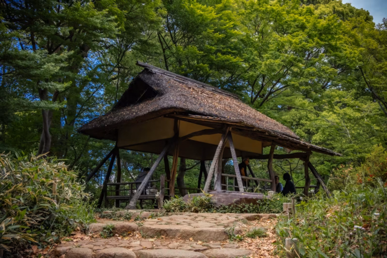 Visitors rest under a rustic thatched-roof teahouse surrounded by lush greenery at Rikugien Gardens.