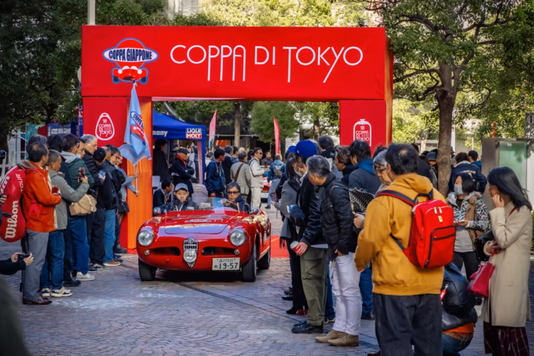 Red classic car drives under a red archway at the Coppa di Tokyo 2024 car rally, surrounded by a crowd of spectators.