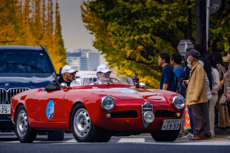 A red vintage Alfa Romeo spider with two occupants drives past spectators on a city street lined with yellow trees.