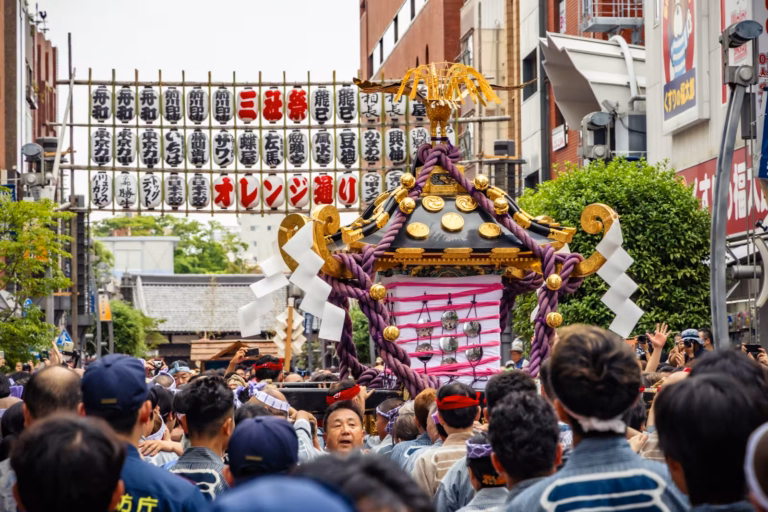 A crowd carries a black and gold mikoshi shrine under a wall of lanterns during Sanja Festival 2025.